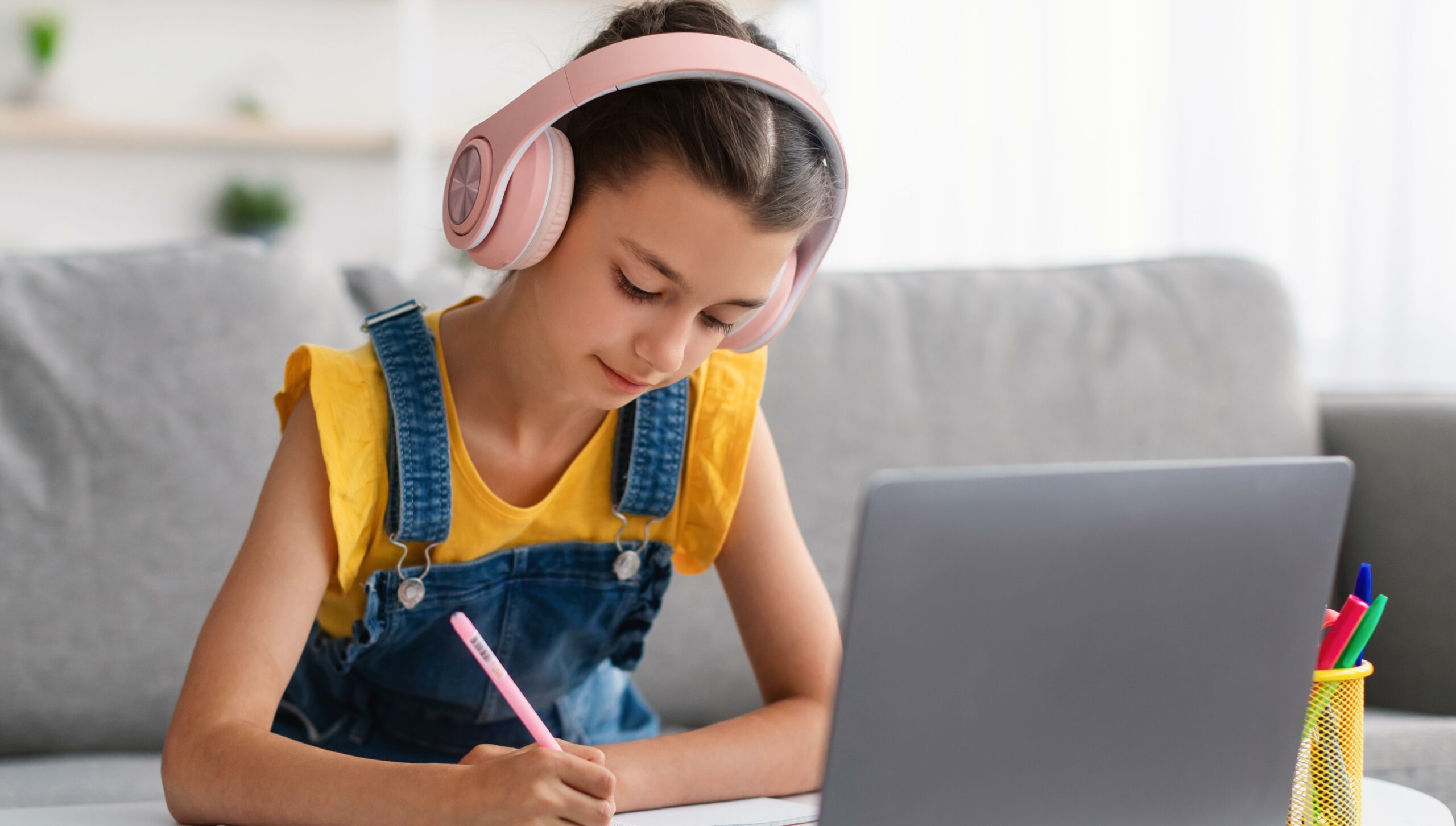 Schoolgirl in pink headset sitting at desk, writing in textbook
