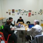 Esta Imagen Muestra A Un Grupo De Personas Sentados En Una Mesa Con Ordenadores Y Documentos Encima De La Mesa.