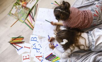 Top view of two tiny girls which are drawing in the coloring book laying on the floor on the blanket Top view of two tiny girls which are drawing in the coloring book laying on the floor on the blanket